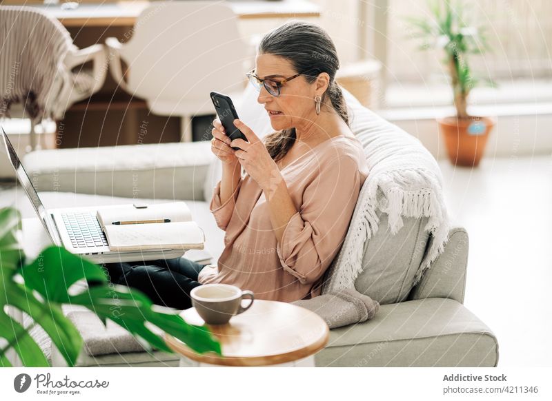 Serious mature woman on mobile phone and working on computer smartphone communicate laptop internet browsing middle age message online sofa room female