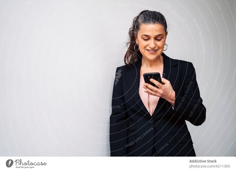 Confident mature woman in formal suit browsing smartphone smile text message entrepreneur happy manager respectable glad business sms cellphone delight