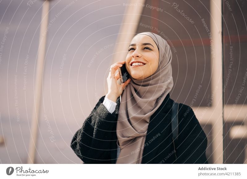 Cheerful Muslim woman speaking on smartphone in city talk hijab smile conversation phone call street female ethnic muslim mobile happy gadget glad content