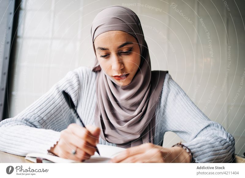 Serious Muslim woman taking notes in notebook in cafe take note freelance work remote write notepad focus female ethnic muslim project thoughtful concentrate
