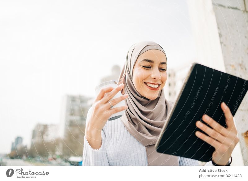 Smiling ethnic woman in hijab using tablet in city browsing takeaway drink smile headscarf street female muslim fountain surfing online internet headdress