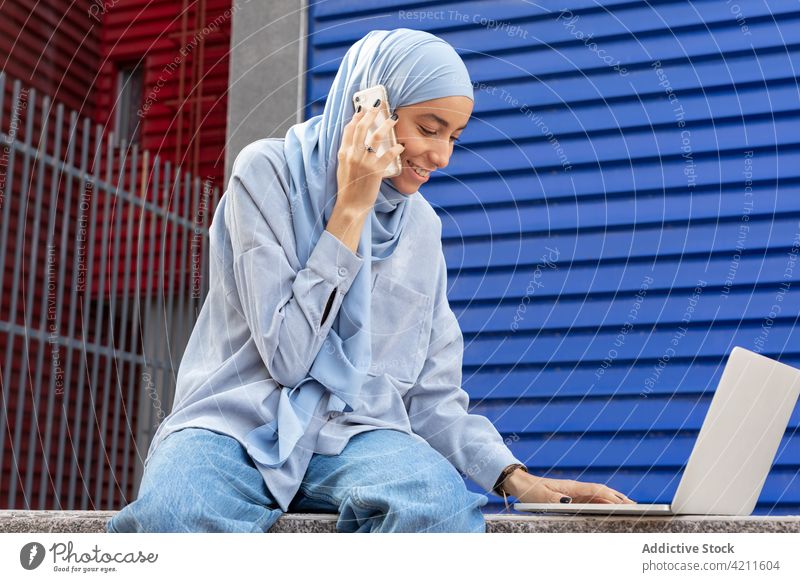 Cheerful ethnic woman with laptop speaking on smartphone in city talk multimedia cheerful pastime urban portrait using gadget device typing content internet