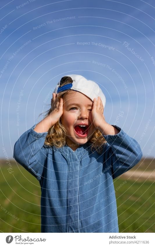 Excited girl in cap in nature summer meadow excited touch head blue sky mouth opened style blond kid happy cheerful pleasure glad positive carefree joy holiday