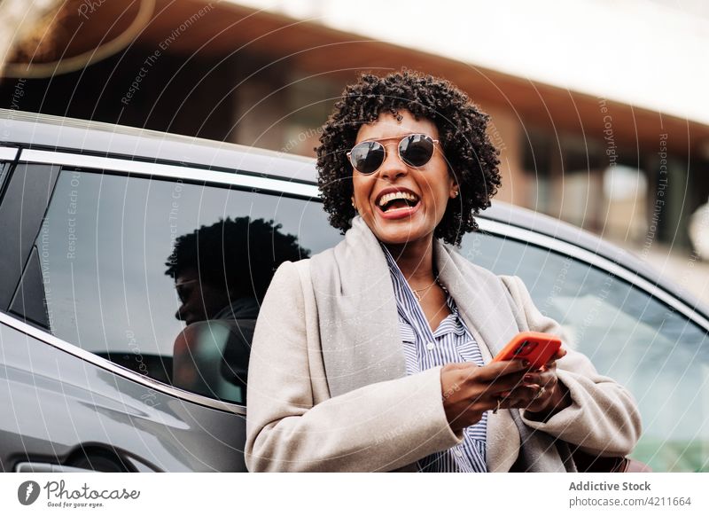 Delighted black woman browsing smartphone near car using laugh happy style delight carefree joy female device cheerful sunglasses mobile gadget vehicle digital