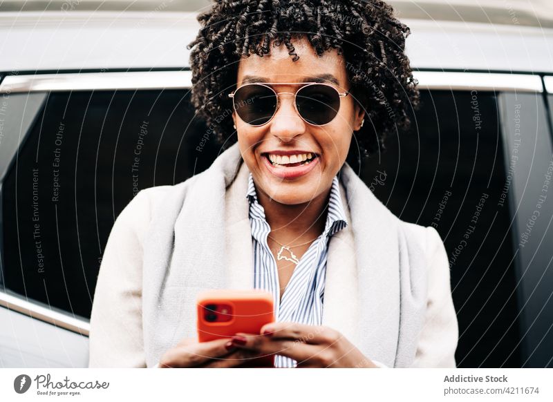 Delighted black woman browsing smartphone near car using laugh happy style delight carefree joy female device cheerful sunglasses mobile gadget vehicle digital