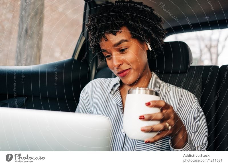 Young black woman with laptop riding in car using coffee morning commute passenger taxi cup cheerful content backseat tws true wireless earbuds thermal elegant