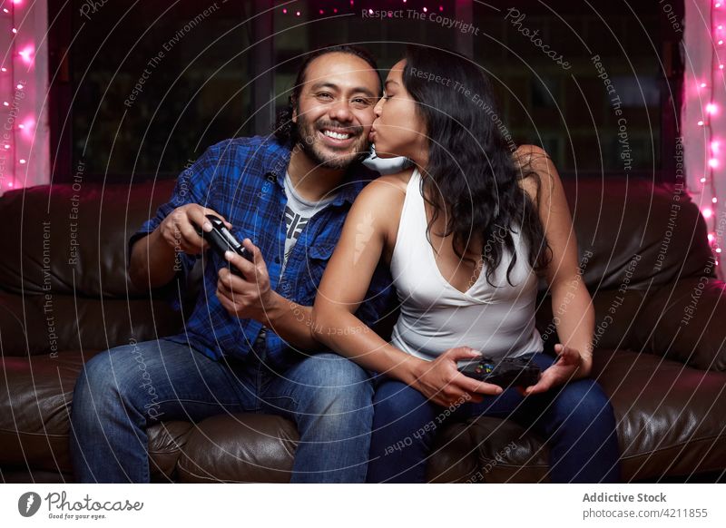 Delighted ethnic couple playing video game together delight videogame kiss excited couch joy pad having fun entertain laugh gamepad free time device pastime