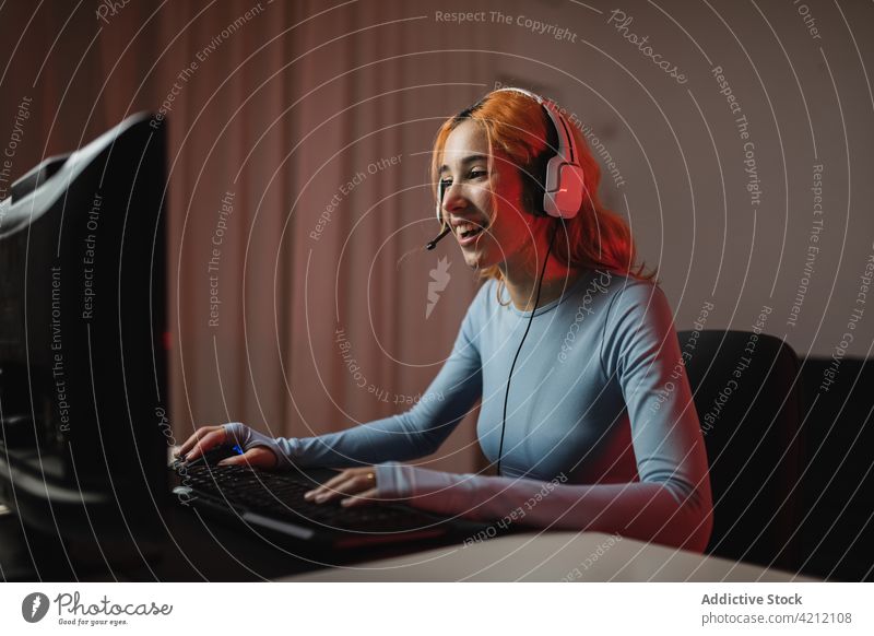 Cheerful gamer playing videogame woman female headphones happy home device gadget using listen glad internet positive entertain online delight cheerful enjoy