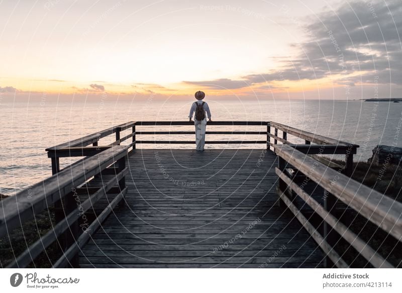 Back view man with backpack and hat standing on a walkway looking at the sunrise near the sea water nature leisure landscape background male sky caucasian