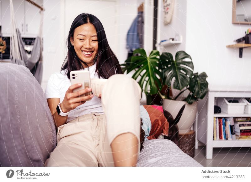 Smiling Asian woman chatting on smartphone on sofa at home text messaging internet online cheerful spare time portrait using gadget device cellphone browsing