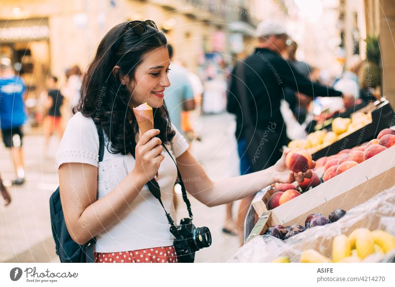 Young tourist woman eating an ice cream and buying some fruit in a street market shopping grocery vacation travel holiday city happy food backpack camera summer