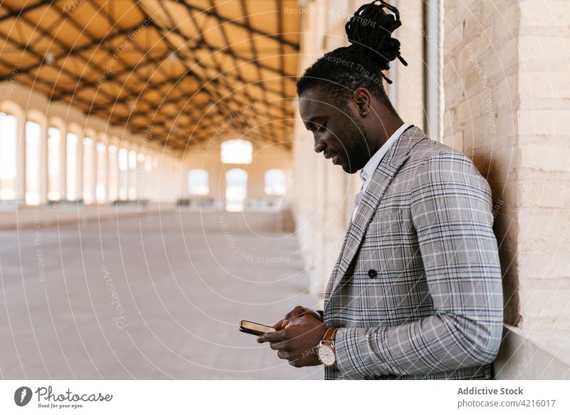 Stylish black businessman chatting on smartphone against building wall stylish individuality dreadlocks internet portrait using gadget online device style