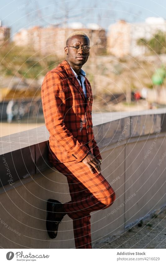 Stylish ethnic man in suit against urban fence fashion style individuality checkered masculine leg raised town tartan portrait eyeglasses cool well dressed