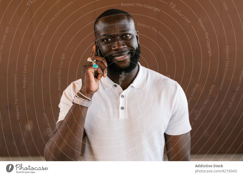 Smiling black man in rings speaking on smartphone send cheerful style phone call accessory talk using gadget device individuality t shirt bracelet smile content