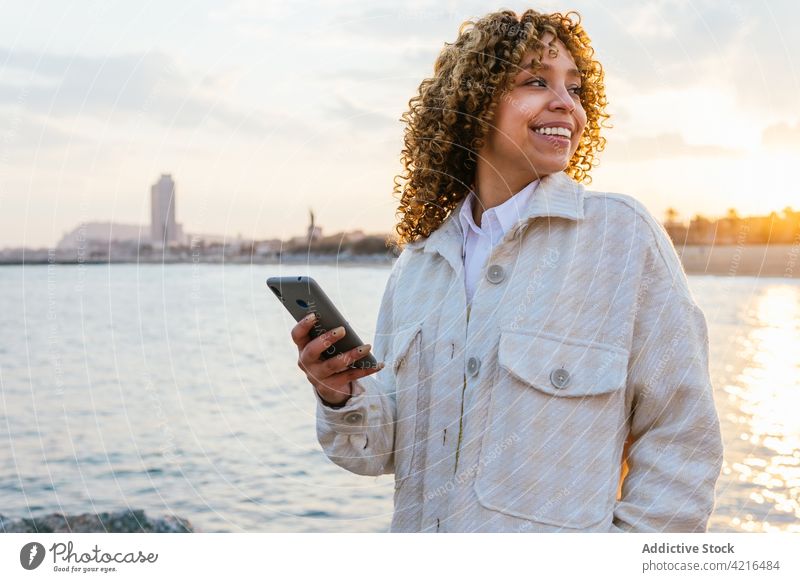 Black woman using mobile phone at seaside seashore sunset arm cheerful female ethnic black african american summer nature smile evening positive holiday