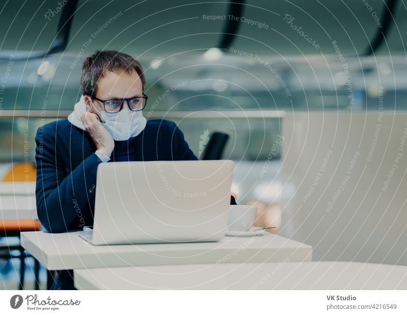 Photo of serious man concentrated in screen of laptop computer, works from distance during coronavirus outbreak, wears protective mask not to spread disease, drinks coffee, sits at white table