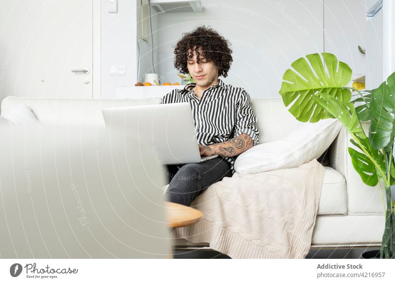 Tattooed man surfing internet on laptop on sofa at home online free time tattoo using gadget device watching coffee cup browsing rest beverage netbook couch