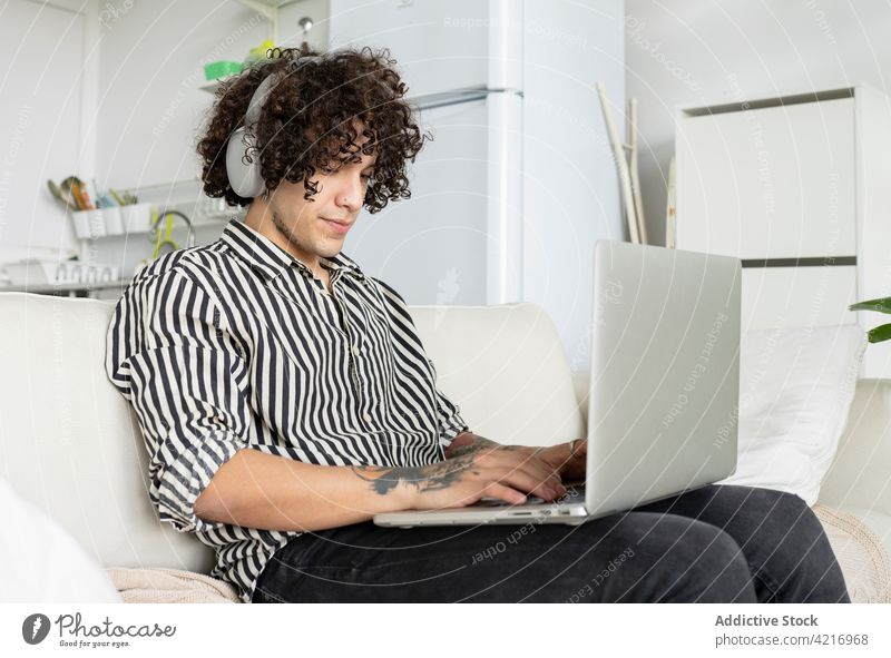 Tattooed man surfing internet on laptop on sofa at home headphones listen online free time tattoo using gadget device watching browsing rest netbook couch