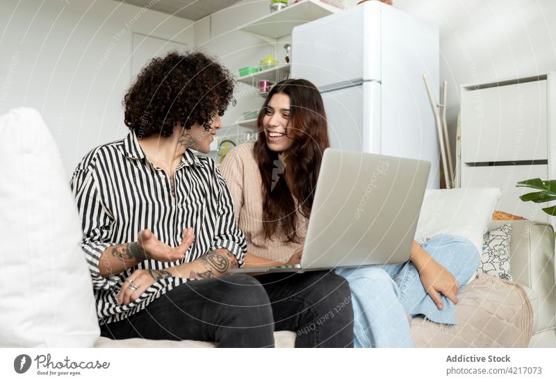 Content couple with laptop speaking on sofa in apartment spend time weekend relationship tattoo sincere home gadget device netbook smile talk hipster content