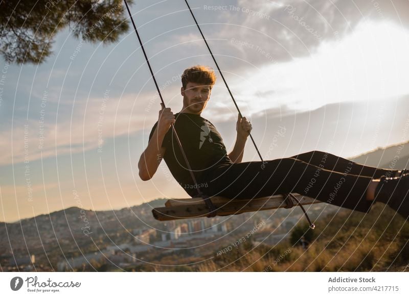 Masculine man on swing under cloudy sky at sunset masculine macho brutal virile mountain sundown town idyllic evening city style casual black apparel sit shiny