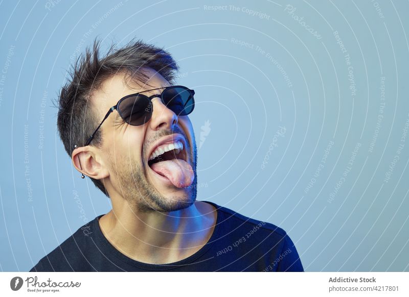 Provocative man showing tongue in studio show tongue tongue out expressive rebel grimace make face eccentric neon light male sunglasses style naughty trendy