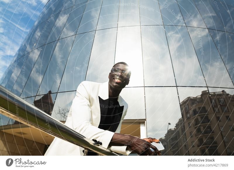 Content black businessman with smartphone on city street toothy smile style friendly candid satisfied reflection portrait cloudy sky enjoy gadget device