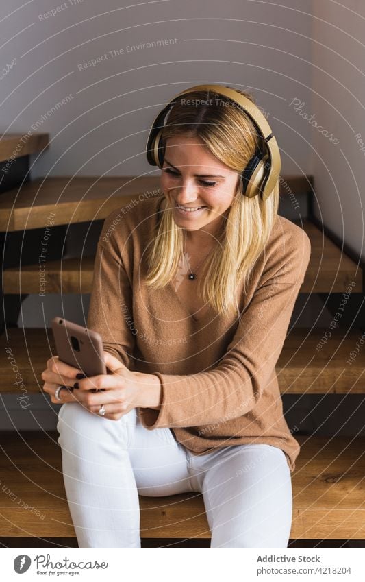 Smiling woman listening to music in headphones at home smartphone browsing song enjoy using female gadget sit surfing device social media chill stair online
