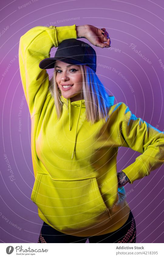 Cool woman in stylish hoodie looking at camera in studio coquette street style smile neon light glow female hat cap outfit cool vivid color blue pink vibrant