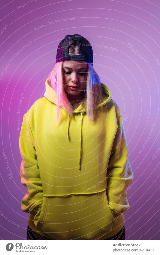 Cool woman in stylish hoodie looking away in studio coquette street style neon light glow female hat cap outfit cool vivid color blue pink vibrant bright