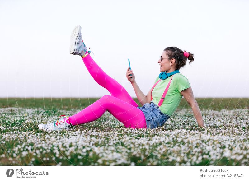 Woman using smartphone and sitting on meadow with legs raised woman lawn countryside joyful carefree gadget outfit browsing positive cheerful content device