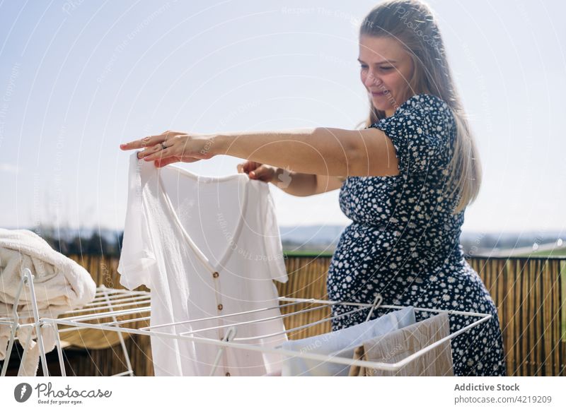 Smiling pregnant woman hanging linen on sunny balcony laundry drying rack smile await motherhood anticipate sky pregnancy tummy maternal cheerful sincere