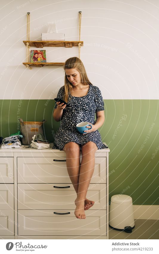 Smiling pregnant woman with tea browsing smartphone at home watching pregnancy sincere prenatal using gadget device phone call await pastime commode sit