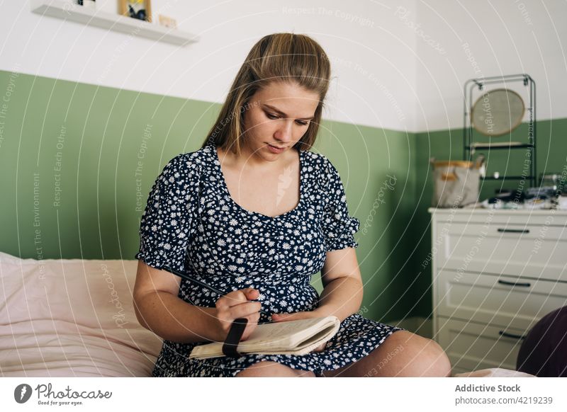 Pregnant woman sitting on bed and writing notes take note notebook attentive feminine gentle house pen write paper notepad ornament cloth home diary room