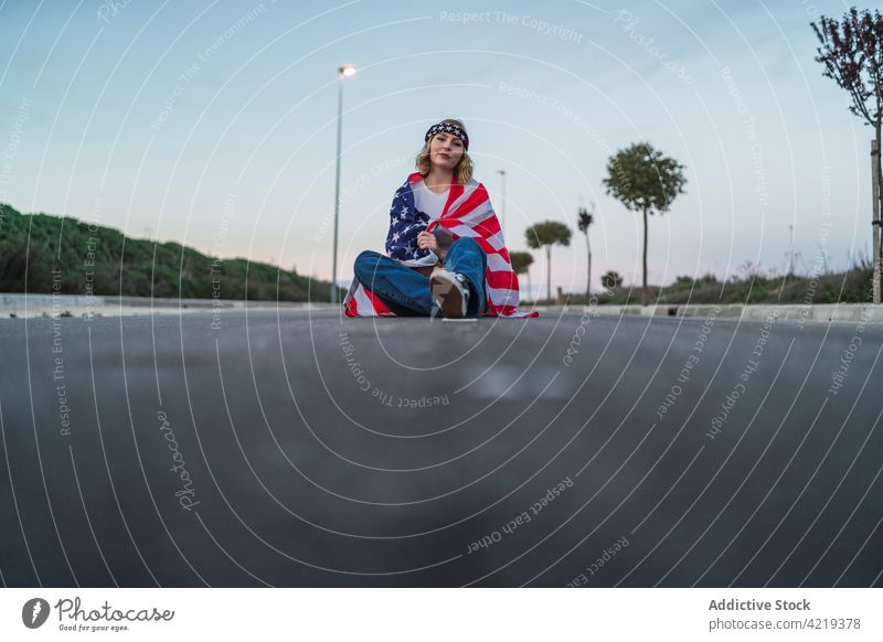 American woman with flag sitting on road patriot carefree wrap national symbol sunset freedom female american usa united states independent 4th july smile pride