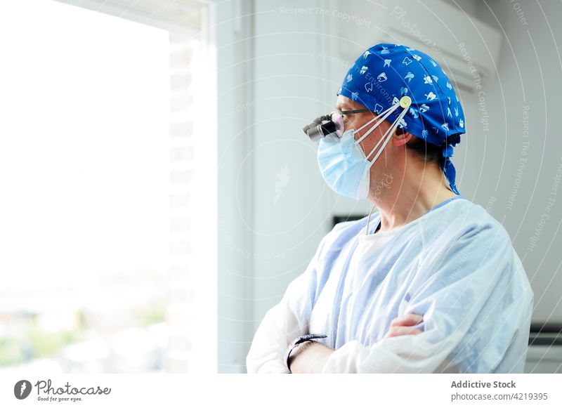Unrecognizable surgeon in medical binoculars with crossed arms in clinic look out window arms crossed uniform professional contemplate man cap ornament cloth