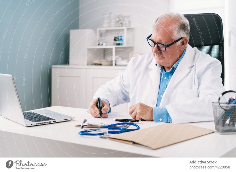 Physician writing on paper at table with tablet in hospital physician take note work professional serious uniform man clinic write sheet elderly gadget device