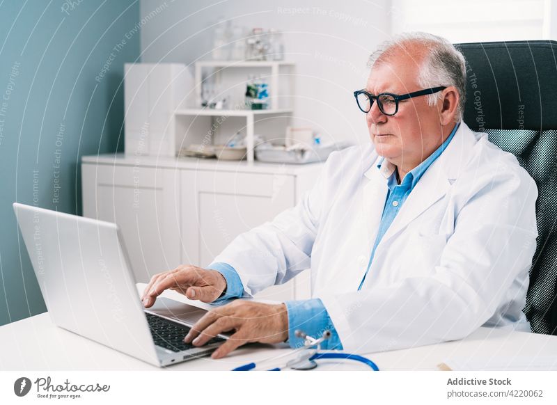 Senior physician working on laptop in hospital doctor typing medical professional job uniform man using gadget device internet serious online surfing workplace