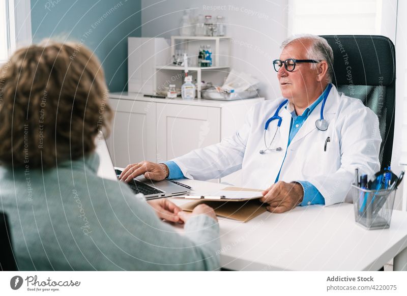 Senior physician using laptop during consultation with anonymous patient doctor typing talk check up health care profession attentive gadget man woman device