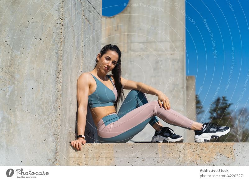 Confident sportswoman resting on concrete surface after workout self assured break body curve sportswear friendly portrait sneakers cement wall blue sky athlete