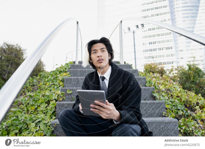 Stylish Asian businessman with tablet on urban stairs internet online style self employed focus city using gadget device shoe sit natural building vegetate