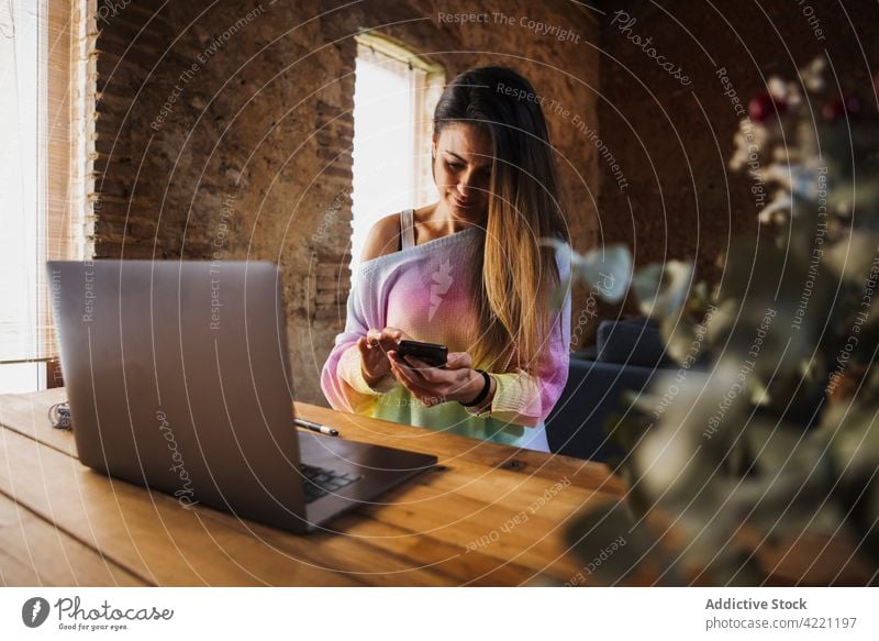 Freelancer chatting on smartphone at table with laptop at home worker freelance self employed loft woman using gadget device text messaging watching employee