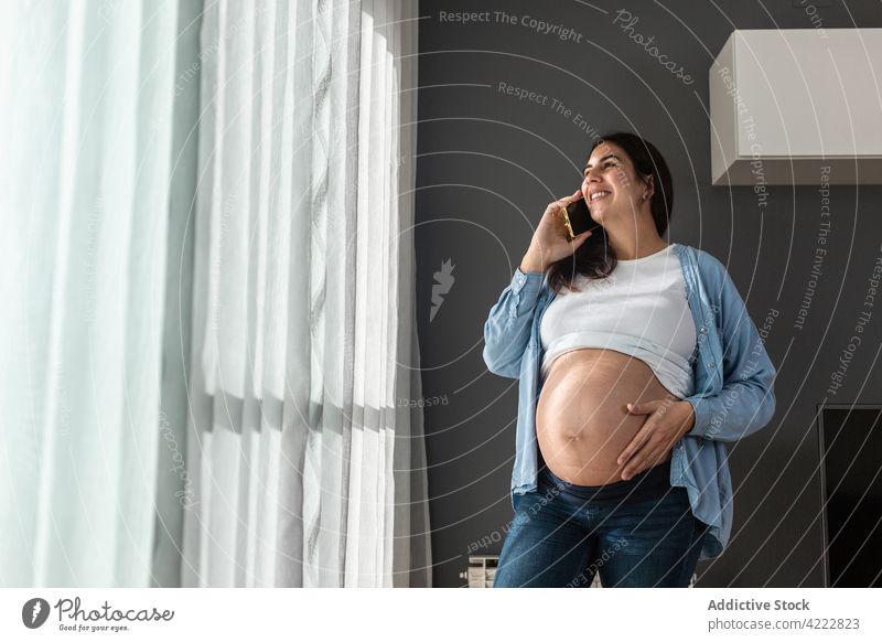 Pregnant woman talking with smartphone at home pregnant speak touch belly social media tummy pregnancy female stand room mobile smile online expect gadget happy