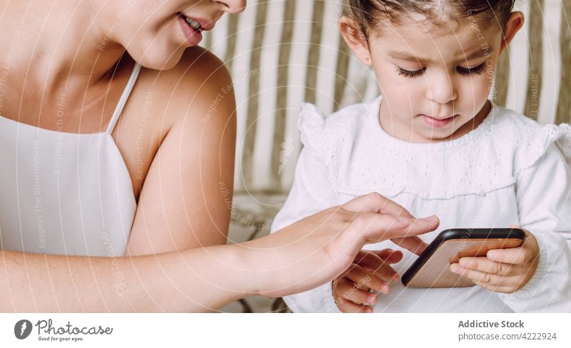 Crop mother and little child using smartphone together daughter help browsing explain ethnic sofa home gadget device online surfing kid motherhood at home