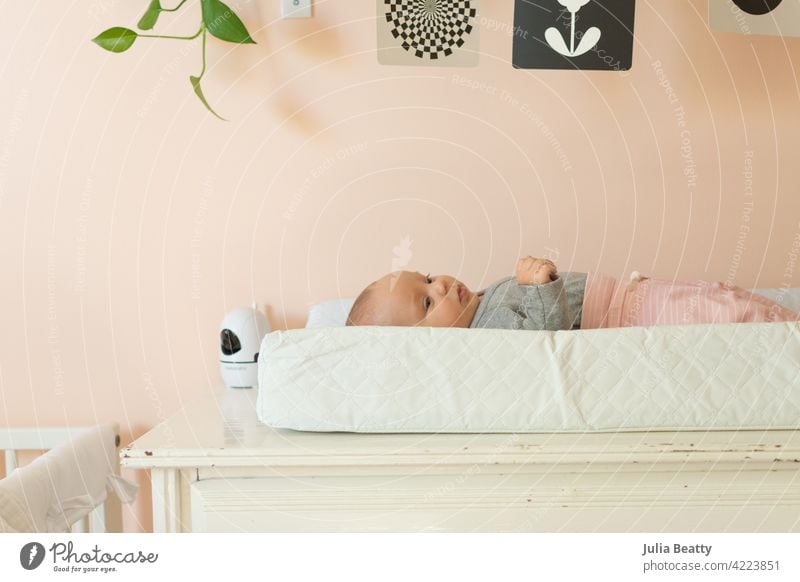 Young baby laying on changing pad in a peach nursery; black and white mobile and baby monitor nearby infant 3 months old 0-6 months old changing table diaper