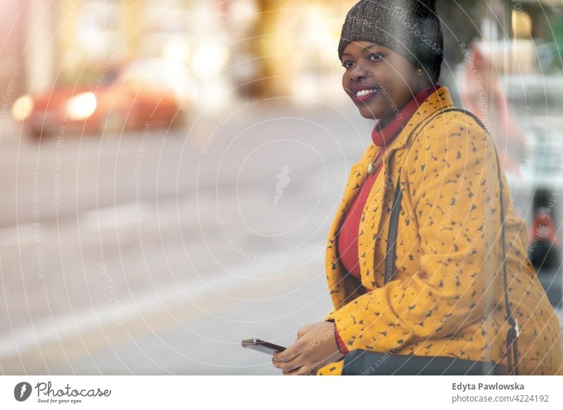 Happy young woman waiting at bus stop holding mobile phone smartphone technology using internet wireless sms text messaging texting outdoors day positivity