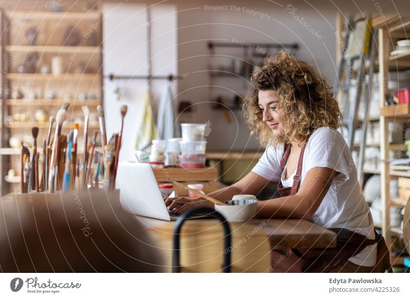 Woman pottery artist using laptop in art studio ceramics work working people woman young adult casual attractive female happy Caucasian enjoying one person