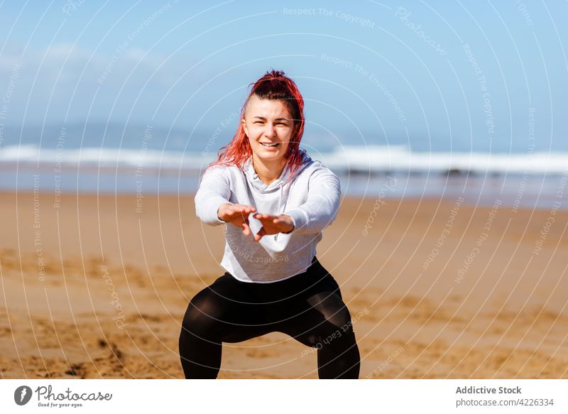 Smiling sportswoman performing squat pose on sea shore exercise training workout energy vitality seashore portrait athlete healthy lifestyle wellness outstretch