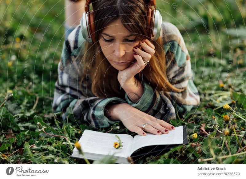 Woman in headphones reading book on grass woman literature knowledge spare time listen using gadget device textbook education music melody millennial attentive