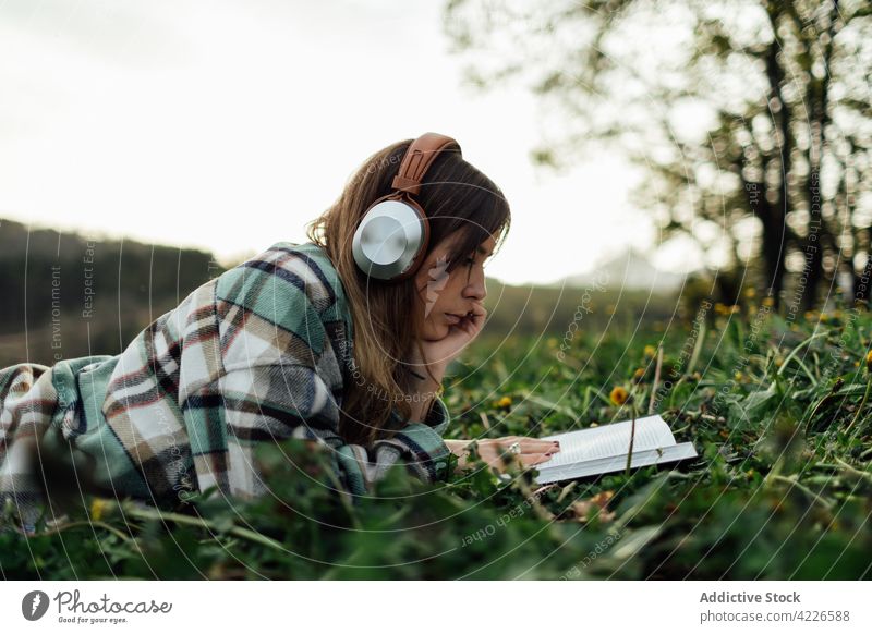 Woman in headphones reading book on grass woman literature knowledge spare time listen using gadget device textbook education music melody millennial attentive