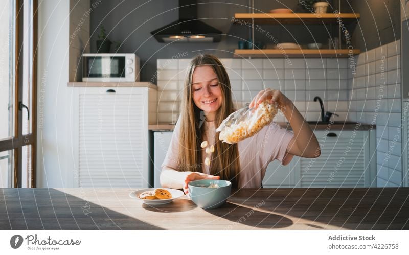 Smiling woman pouring corn rings into bowl in kitchen cereal breakfast prepare cookie biscuit sweet smile baked table domestic cheerful bag flow oatmeal